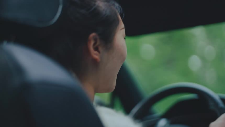 A woman in the drivers seat of a car smiling and appearing to be enjoying the drive she briefly turns her head to look out the side window with a joyful expression