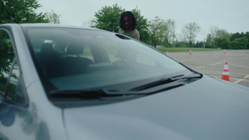 A grey car is parked in a lot lined with orange traffic cones as two people talk in front of the car