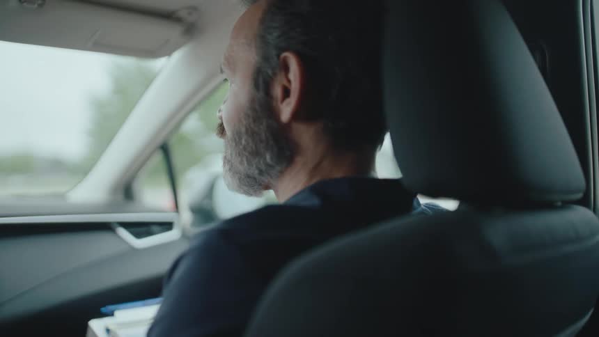 A bearded man is seated in the passenger seat of a car looking out the window he appears to be talking or gesturing with his right hand as if in conversation while the car seems to be in motion