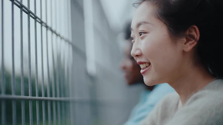 Two women one chinese and one black sit close to a fence talking and smiling as a light breeze gently blows through their hair at midday