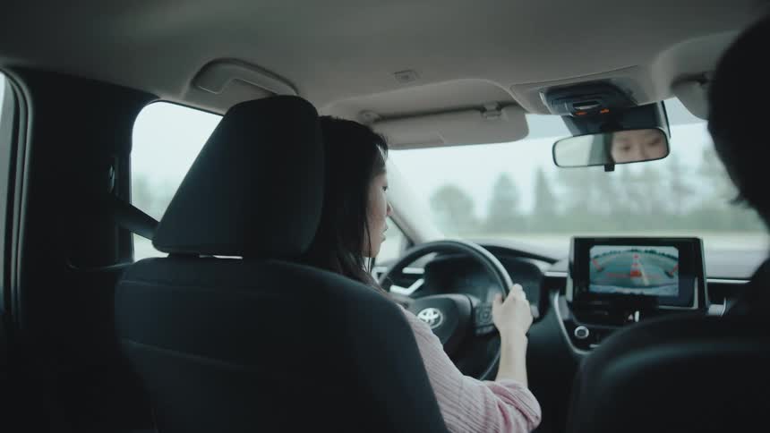A student driver with dark hair sits in the drivers seat of a car on a driving course as viewed from the back passenger seat with a rearview camera displaying a red safety outline