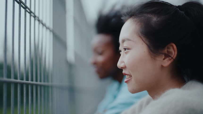 A side profile of a young woman behind a fence looking ahead and smiling another person is visible in the background also looking in the same direction