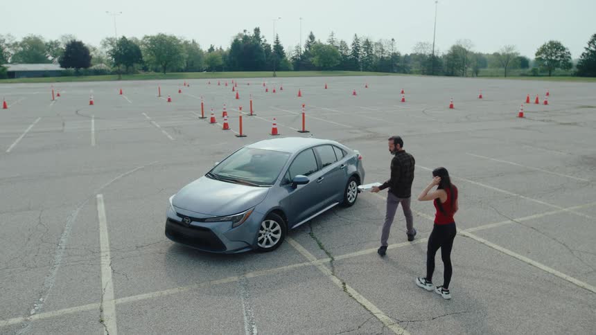 Two people stand near a car in an empty lot set up for a driving test with traffic cones