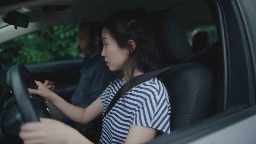A young woman in the drivers seat of a car wearing a blue and white striped shirt and a seatbelt with her hands on the steering wheel an older man with a beard is visible in the passenger seat appearing to be observing or instructing her