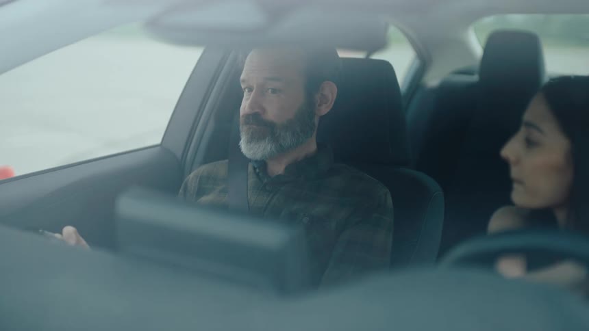 A closeup from inside a car shows a bearded man in the passenger seat intently looking out the window his head slightly turned beside him a woman is partially visible in the drivers seat suggesting a shared journey or conversation