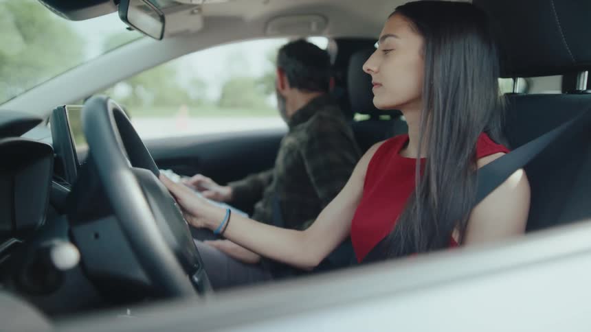 A young woman with long dark hair sits in the drivers seat of a car attentively holding the steering wheel indicating she is either driving or about to beside her a man in the passenger seat is focused on a laptop suggesting they are on a journey