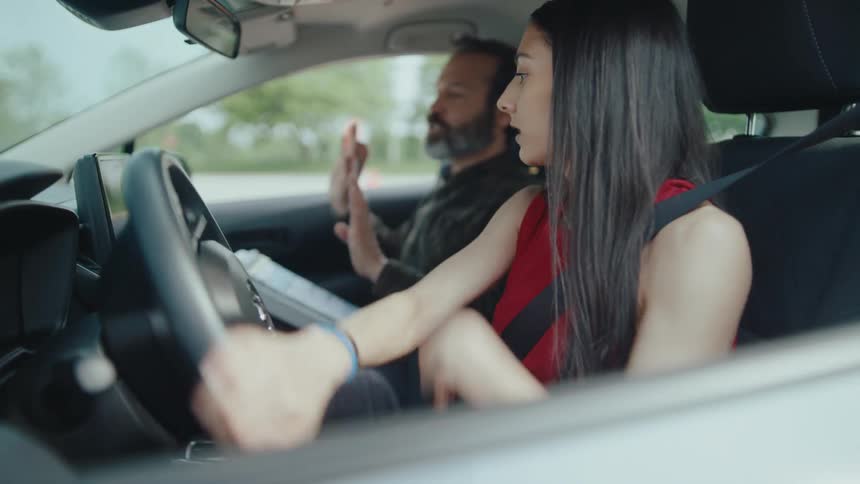 A young woman drives in a parking lot with a driving instructor beside her moving back and forth as directed by the instructor at midday