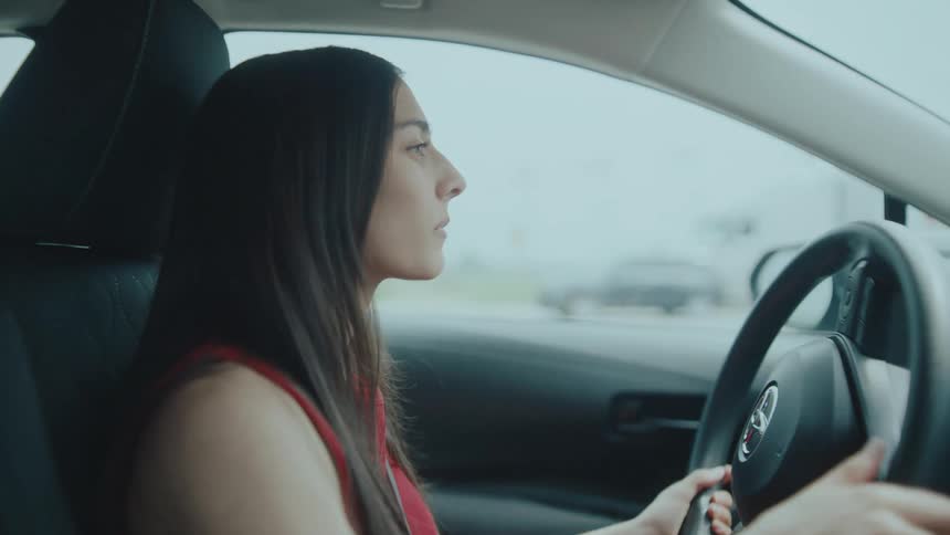 A woman drives a car while gripping the steering wheel with both hands in a city street