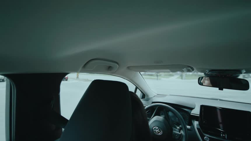 Seen from the back seat a woman drives a modern car making a turn on a road as her face is reflected in the rearview mirror