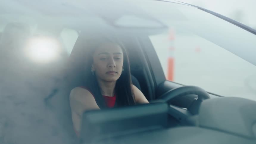 A woman with long dark hair visible through the windshield of a car appearing distressed and running her hand through her hair the car seems to be stopped possibly in traffic as she looks around with a frustrated expression