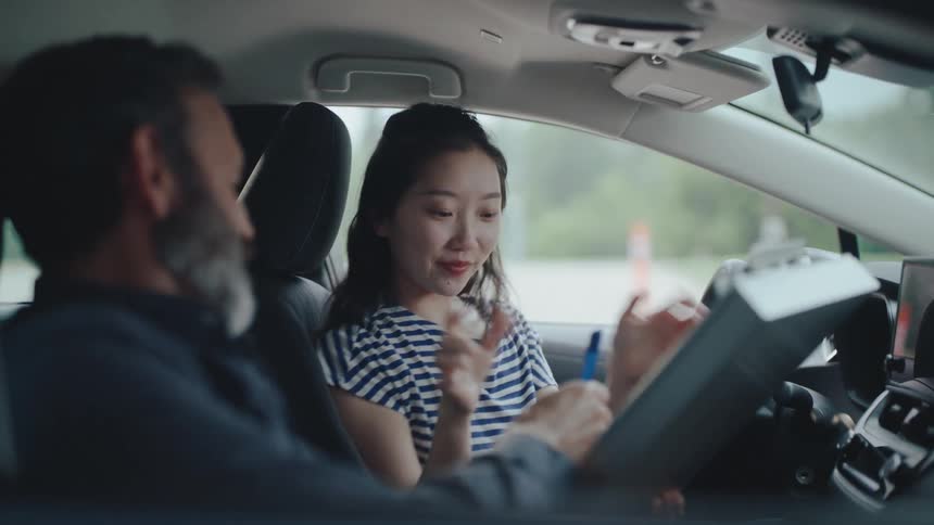 A bearded man sits in the passenger seat of a car watching a woman in the drivers seat as she smiles and adjusts the steering wheel the scene suggests a driving lesson or a shared car journey between the two individuals