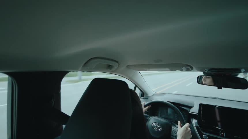 A view from the back seat shows a woman driving a car down the road with her reflection visible in the rearview mirror