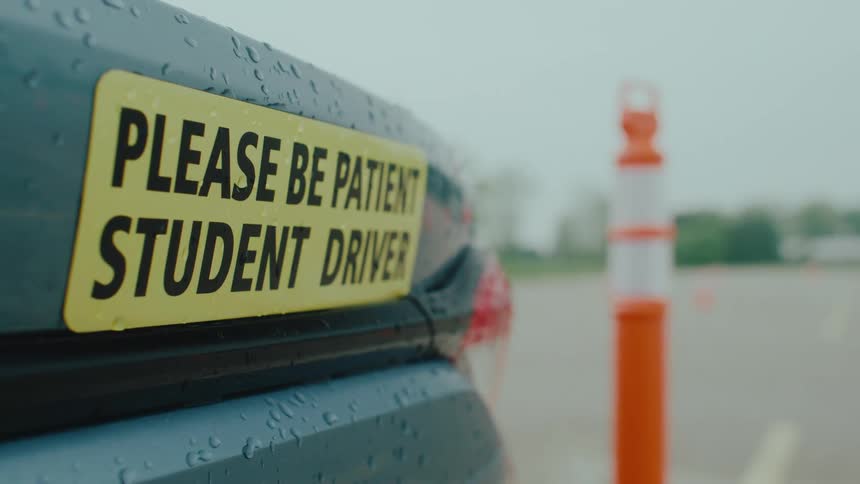 A yellow please be patient student driver magnet is prominently displayed on the wet trunk of a car with a blurry orange traffic cone in the background the scene clearly indicates a vehicle being used for driving instruction