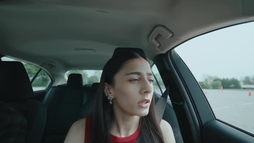 A woman in a red top sits in the drivers seat looking out the window thoughtfully