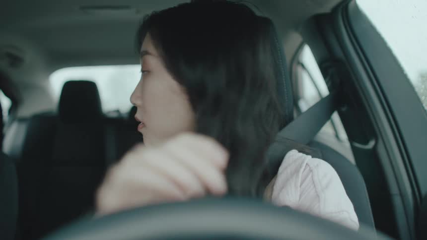 A woman in the drivers seat of a car wearing a lightcolored top and a seatbelt she looks around cautiously and appears focused on driving or parking the car
