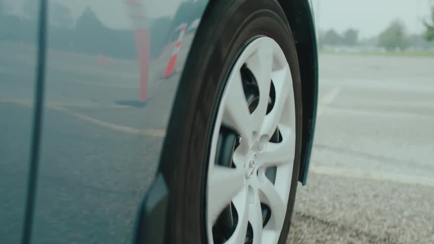 The right wheel of a car moves forward with reflections of traffic cones visible on the cars body as it drives through a public parking area at midday