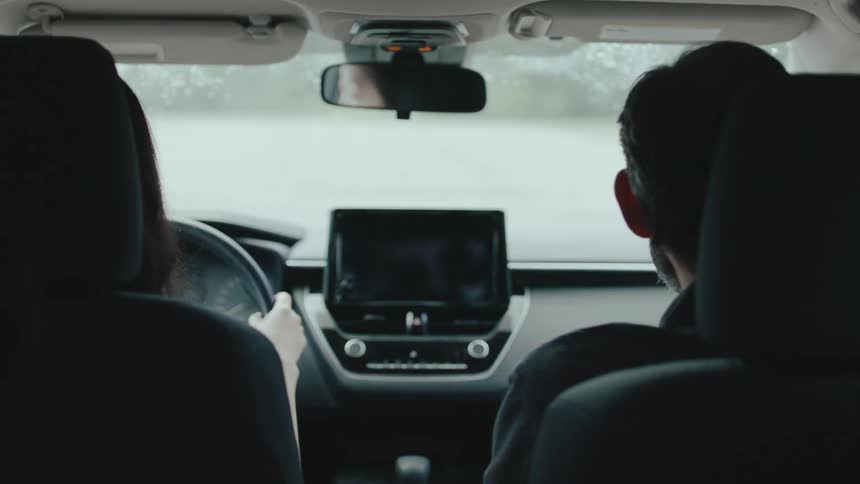 A man and a woman are sitting in the front seats of a car and the man is pointing to his side while talking to her