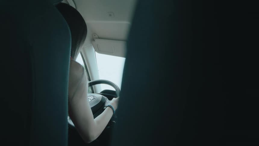 A woman with dark hair in the drivers seat of a car as viewed from the back seat she is holding the steering wheel and her arm with a bracelet is visible as she focuses on driving