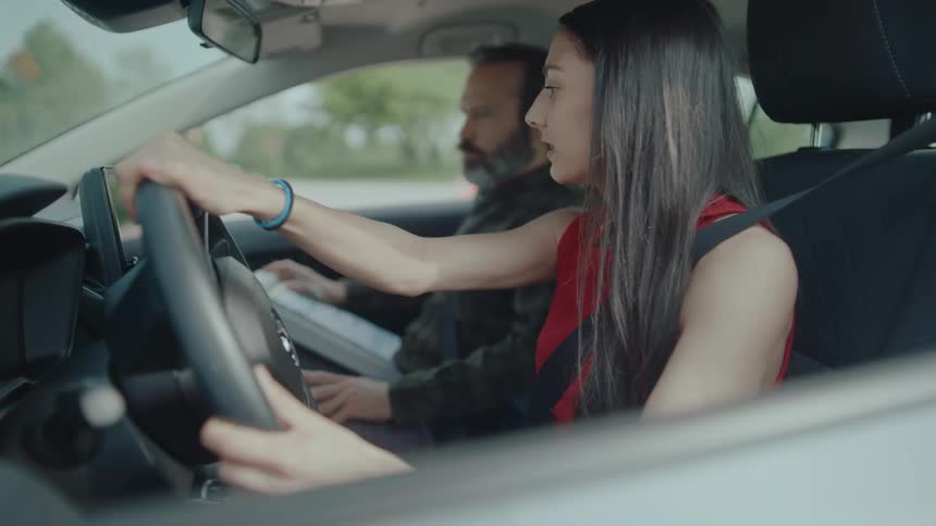 A young woman with long dark hair behind the wheel of a car with a bearded man in the passenger seat they appear to be in a driving lesson or a similar instructional setting as the woman maintains a focused expression