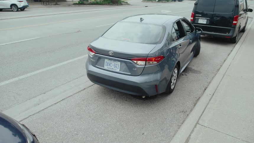 A gray sedan parked on the side of a road next to a black van cars are visible driving past on the main road in the background