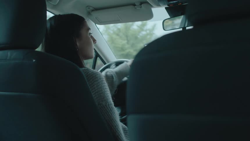 A view from the back seat of a car focusing on the driver who has long dark hair and is wearing a lightcolored sweater she is actively driving with her hands on the steering wheel and occasionally looks to her left as if checking her blind spot
