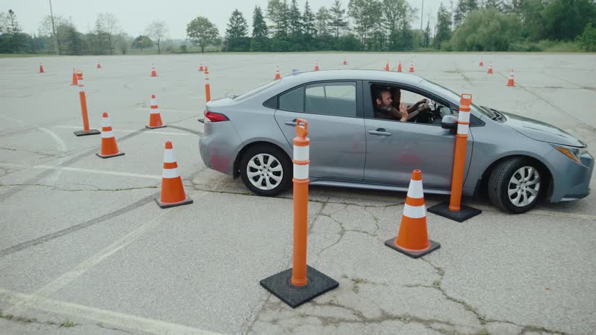 A silver sedan is shown navigating a driving course marked by numerous orange traffic cones suggesting a driving test or practice session the car slowly maneuvers around the cones with a driver visible inside demonstrating careful control