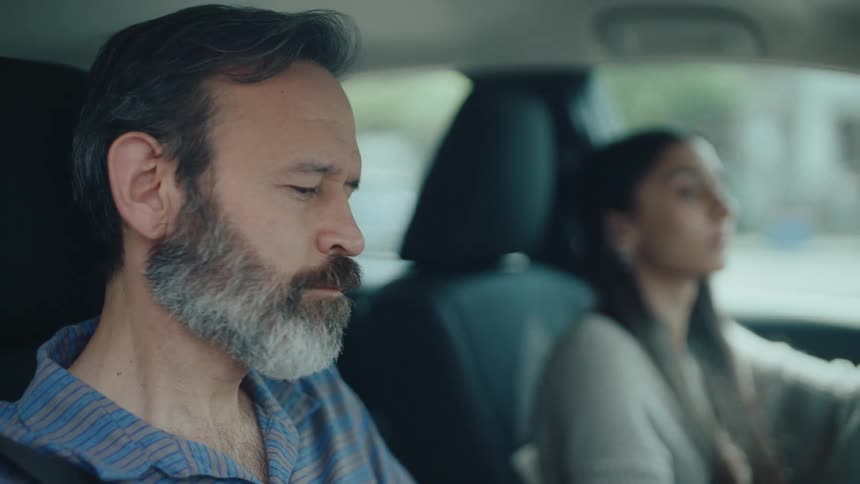 A man with a grey beard in the passenger seat of a car looking forward with a serious expression in the blurred background a woman is visible in the drivers seat suggesting they are a couple on a car journey