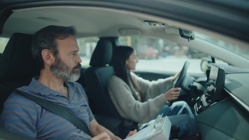 An instructor observes and takes notes while a female student driver navigates a car during a drivers test