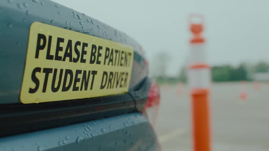 A car with a please be patient student driver sticker is driving in a driving course