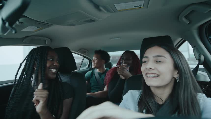 A group of four happy friends laugh and talk together while driving in a car emerging from a dark garage into the daylight