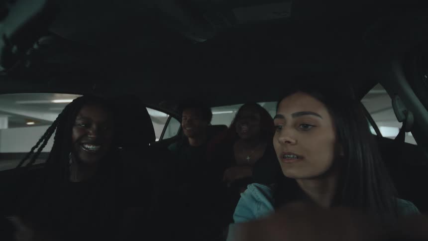 Four young adults three women and one man inside a car the two women in the front seats are looking forward while the two in the back appear to be talking or interacting with each other