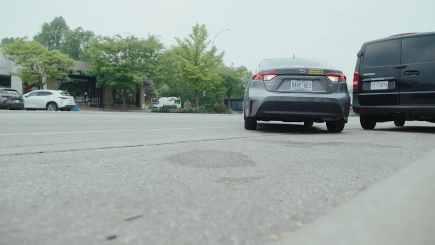 A gray sedan driving past the camera and then parking on the side of the road next to a black van in the background other cars are seen driving on a wider road and a commercial building with trees is visible