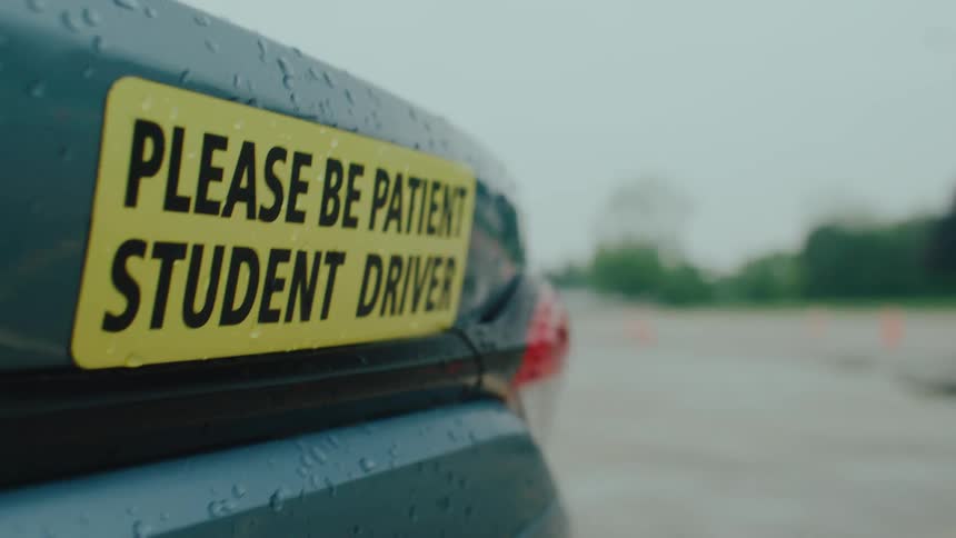 A bright yellow sign reading please be patient student driver is attached to the back of a wet dark teal car on a driving course