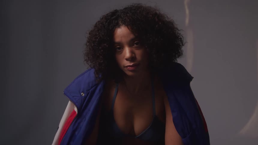 A woman with curly hair wearing a blue and red vest over a dark top she looks directly at the camera with a confident and serious expression