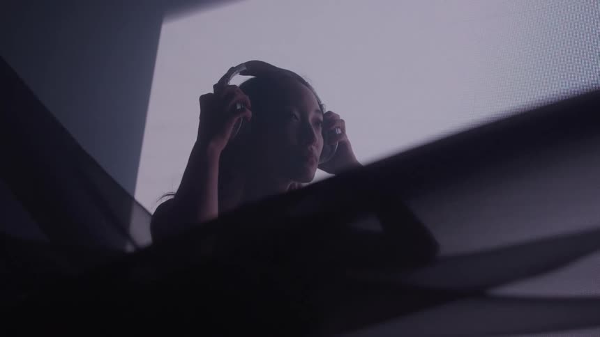 An asian woman is placing a pair of headphones on her ears while standing in front of a projector screen