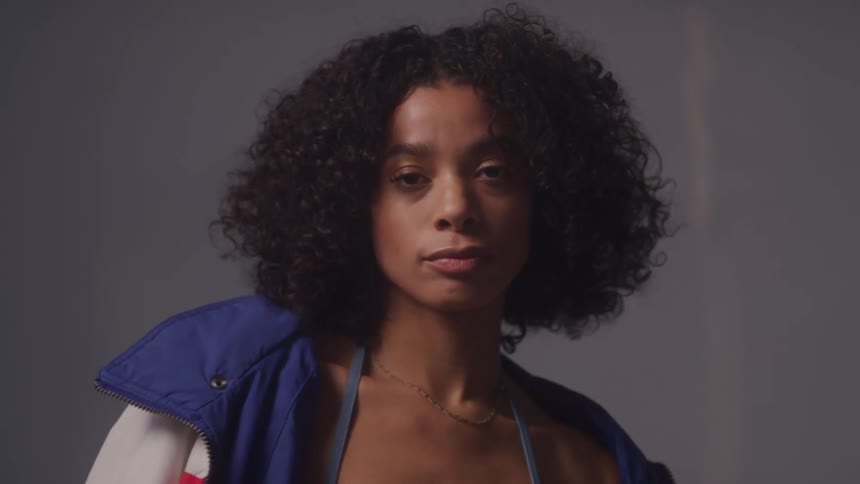 A woman with curly hair wearing a blue and red vest over a dark top she looks directly at the camera with a confident and serious expression then adjusts her hair and neck