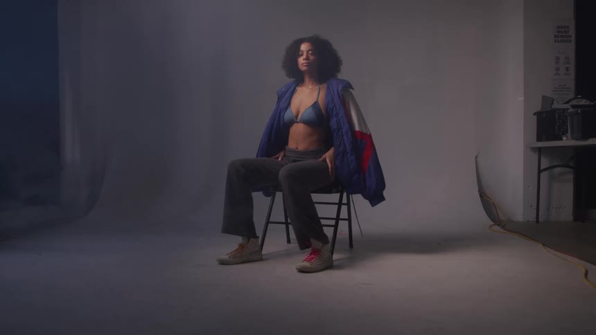 A woman with dark curly hair sits on a black chair models wearing a light blue bra dark pants and a blue red and white jacket draped over her shoulders in a room with a white backdrop