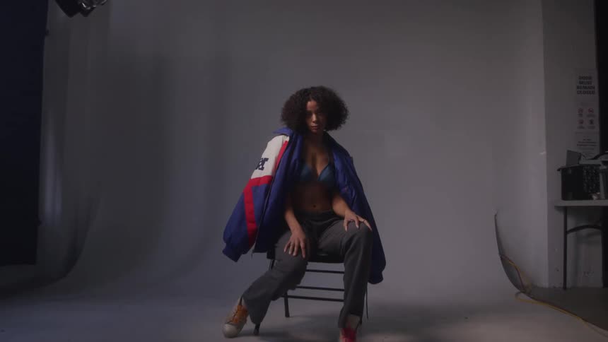 A black model poses confidently in a studio wearing an open jacket bra and pants