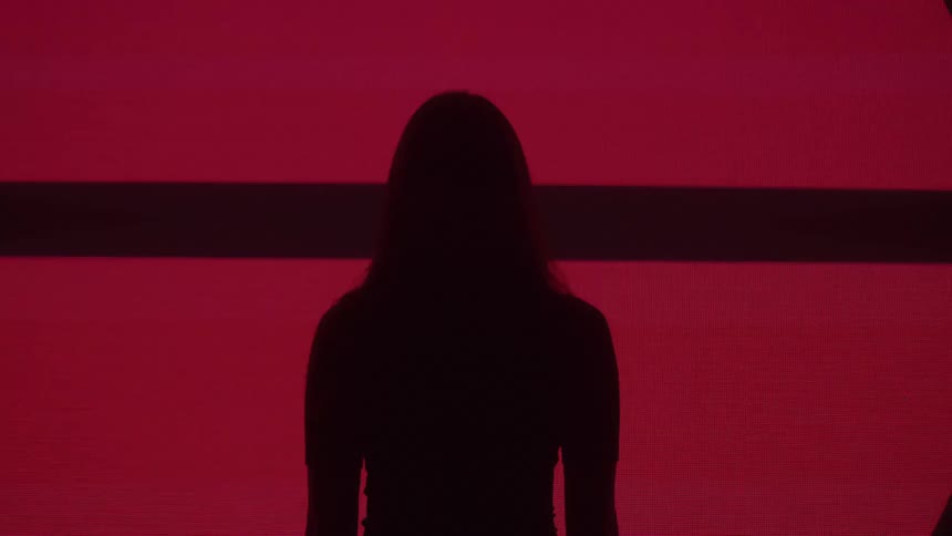 A silhouetted woman stands before a large pink screen displaying a rapid succession of bold dark symbols and patterns