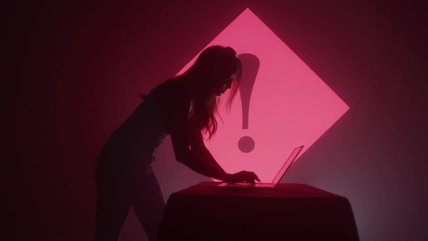 A woman operates a laptop in a club with a screen beside her displaying various red signs while the atmosphere remains dark