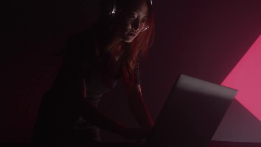 A woman wearing headphones is illuminated by the glow of her laptop screen in a dimly lit room her focused expression suggests she is deeply engaged with her work possibly late at night