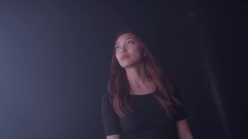 A person stands in a dimly lit room illuminated by a strong light source entering from the left highlighting their features and casting soft shadows the individual looks upwards and around seemingly observing or interacting with the light