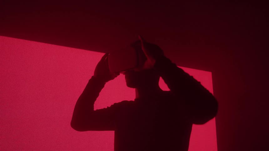 Close up silhouette of a young man putting on vr goggles while standing in a red lit room