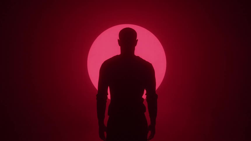 A dark silhouette of a person standing against a red background behind the silhouette a neon red outline of a person appears and then transforms into a square shape the word filmsupply is faintly visible across the persons torso