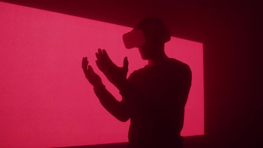 Silhouette of a young man standing in a red lit room while playing a virtual reality game with vr goggles