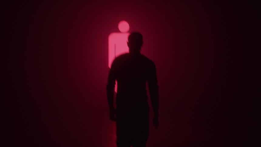 The silhouette of a person walking towards a light with glowing red symbols in a dark indoor environment
