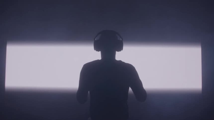 A silhouetted figure stands in the dark and puts on headphones as a bright horizontal light expands behind them creating a powerful backlit effect