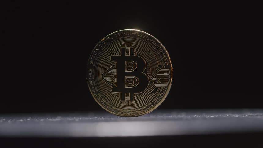 A shimmering golden bitcoin centrally placed on a dark surface spins and then comes to rest reflecting light dramatically its highly detailed surface captures the eye emphasizing its metallic gleam and intricate design