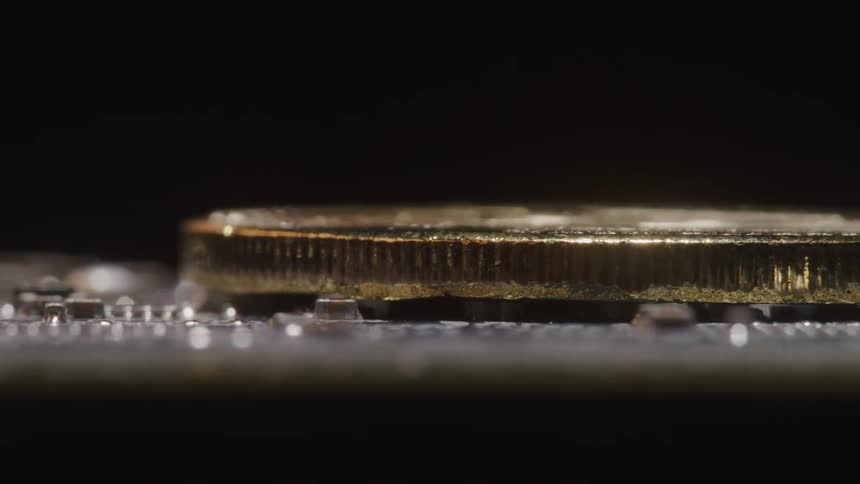 A golden colored metal coin rests on its face atop a printed circuit board
