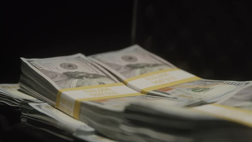 A closeup rack focus shot shifts between stacks of onehundreddollar bills bundled in currency straps on a dark surface
